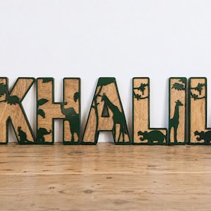 May include: A set of wooden letters spelling the name KHALIL, featuring a layered design with a dark green forest-themed overlay and a natural wood grain background. Each letter incorporates intricate cutouts of various safari animals.