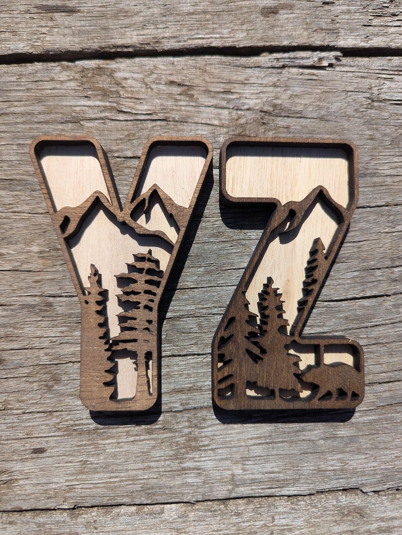 May include: Two wooden letters Y and Z featuring a layered cutout design. The dark brown outer frames enclose a light wood background depicting a mountain range, evergreen trees, and a small bear silhouette at the base of the letter Z.