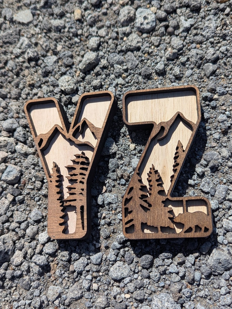 May include: Two wooden letters Y and Z featuring a layered design. The dark brown outer frame contains a light tan wood inlay depicting a mountain range, evergreen trees, and a bear silhouette within the letter Z, creating a dimensional effect.