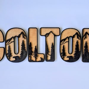 May include: A set of wooden letters spelling the name COLTON, featuring a layered design with a light wood finish and black cutouts of mountain ranges, pine trees, and small bear silhouettes integrated into each individual letter shape.
