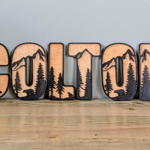 May include: A set of wooden letters spelling the name COLTON, featuring a layered design with a natural light wood finish on the front and a dark, laser-cut silhouette of mountains, pine trees, and bears integrated into each letter.