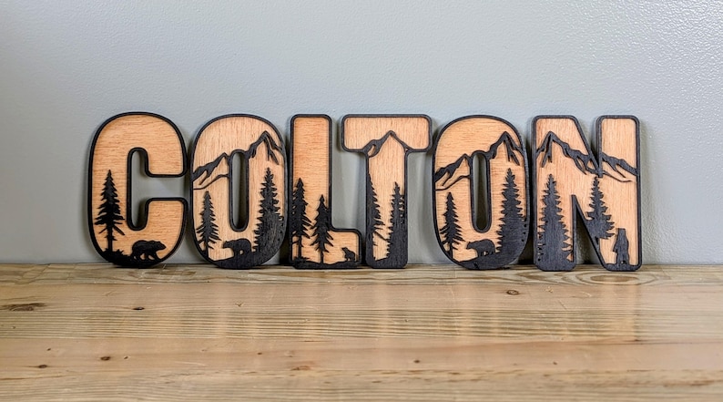 May include: A set of wooden letters spelling the name COLTON, featuring a layered design with a natural light wood finish on the front and a dark, laser-cut silhouette of mountains, pine trees, and bears integrated into each letter.
