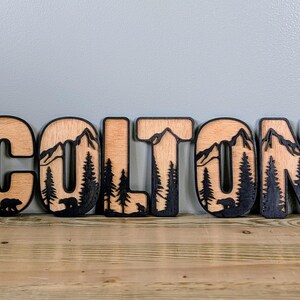 May include: A set of six wooden letters spelling the name COLTON, featuring a layered design with a natural wood finish and black cutouts depicting mountain peaks, evergreen trees, and small bear silhouettes within each individual letter.