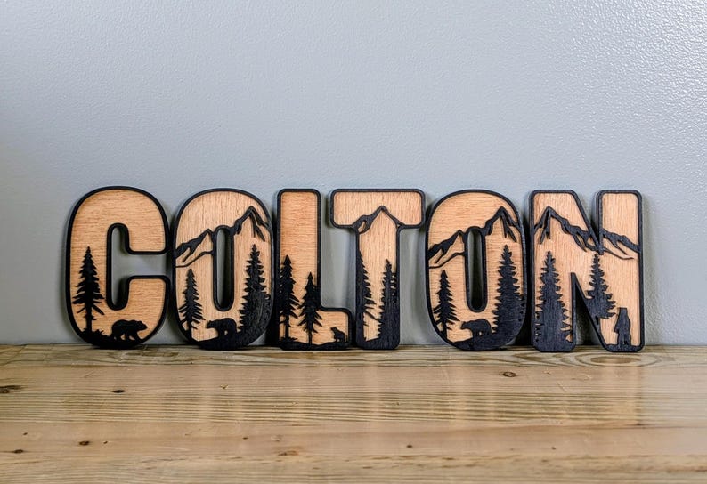 May include: A set of six wooden letters spelling the name COLTON, featuring a layered design with a natural wood finish and black cutouts depicting mountain peaks, evergreen trees, and small bear silhouettes within each individual letter.