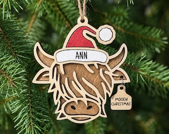 May include: A wooden Highland cow ornament featuring a red and white Santa hat with the name ANN printed on the brim. A small wooden tag attached to the side reads MOOEY CHRISTMAS. The ornament hangs from a brown twine string.