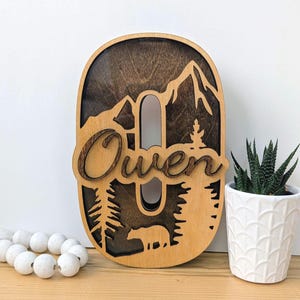May include: A wooden letter O sign featuring a layered mountain and forest landscape with a bear silhouette. The name Owen is written in cursive script across the center. The piece has a dark stained background and a light wood front layer.