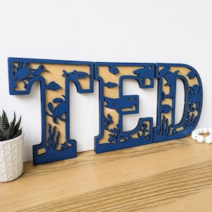 May include: Three dark blue wooden letters spelling TED, featuring intricate cutouts of marine life including sharks, crabs, fish, an octopus, and seaweed. The letters have a natural wood-colored backing visible through the detailed ocean-themed silhouettes.