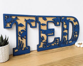 Peut inclure: Trois lettres en bois bleu foncé formant le mot TED, avec des découpes complexes représentant la vie marine, notamment des requins, des crabes, des poissons, une pieuvre et des algues. Le fond en bois naturel est visible à travers les silhouettes.
