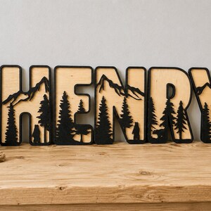 May include: A set of five wooden letters spelling the name HENRY, featuring a layered design with a light wood background and black cutouts depicting mountain peaks, pine trees, and bear silhouettes integrated into each letter shape.