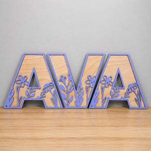 Könnte beinhalten: Ein Holzschild mit dem Namen AVA in Großbuchstaben. Jeder Buchstabe hat einen hellvioletten Rand und eine Aussparung aus Naturholz. Die Buchstaben sind mit filigranen, lasergeschnittenen Blumen- und Blattmustern in Hellviolett verziert.