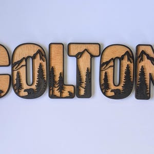 May include: A set of six wooden letters spelling the name COLTON. Each letter features a layered design with a light wood grain background and a dark brown foreground cutout depicting mountain peaks, evergreen trees, and small bear silhouettes.