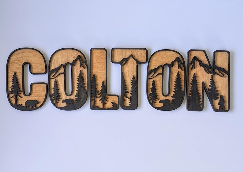 May include: A set of six wooden letters spelling the name COLTON. Each letter features a layered design with a light wood grain background and a dark brown foreground cutout depicting mountain peaks, evergreen trees, and small bear silhouettes.