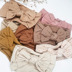 May include: A collection of eight knotted headbands in various neutral colors, including beige, brown, and pink. The headbands are made of a soft, textured fabric and feature a large, knotted bow.