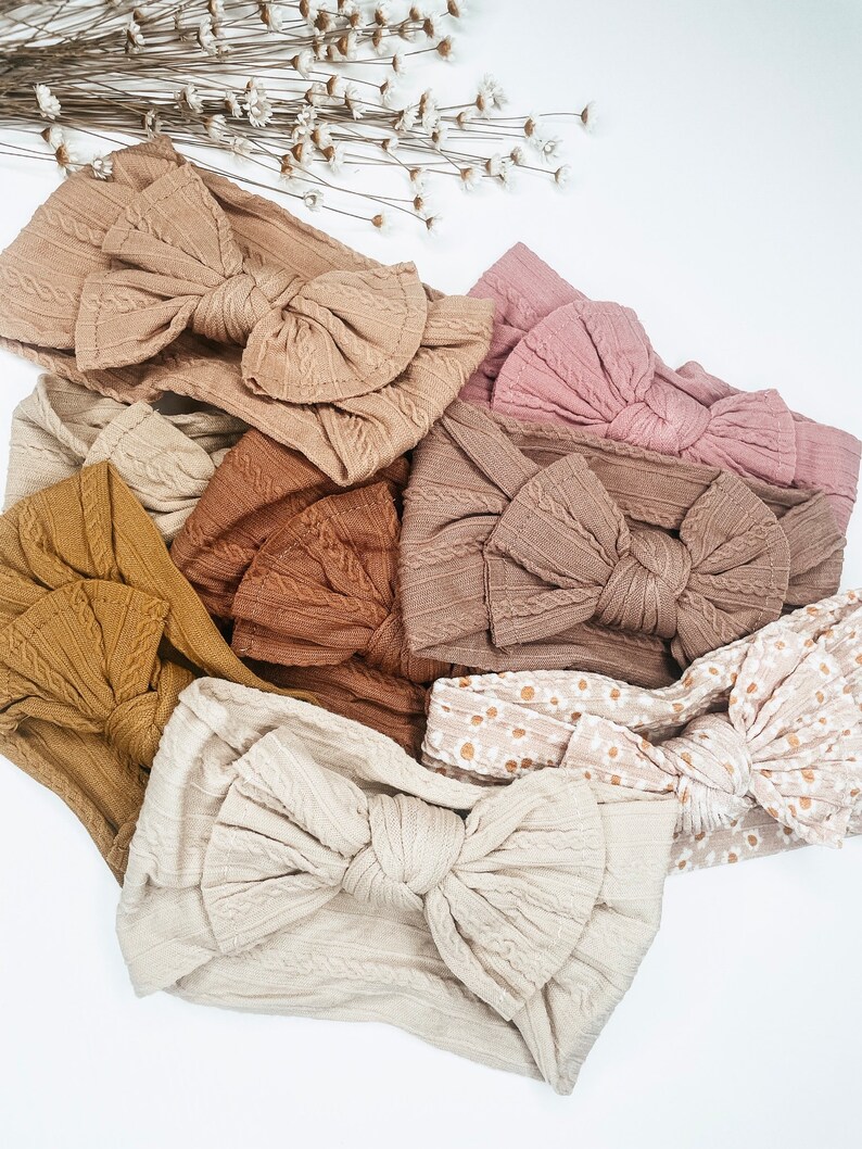 May include: A collection of eight knotted headbands in various neutral colors, including beige, brown, and pink. The headbands are made of a soft, textured fabric and feature a large, knotted bow.
