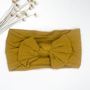 May include: A mustard yellow knotted headband with a cable knit texture. The headband is made of soft, stretchy fabric and is perfect for adding a touch of style to any outfit.