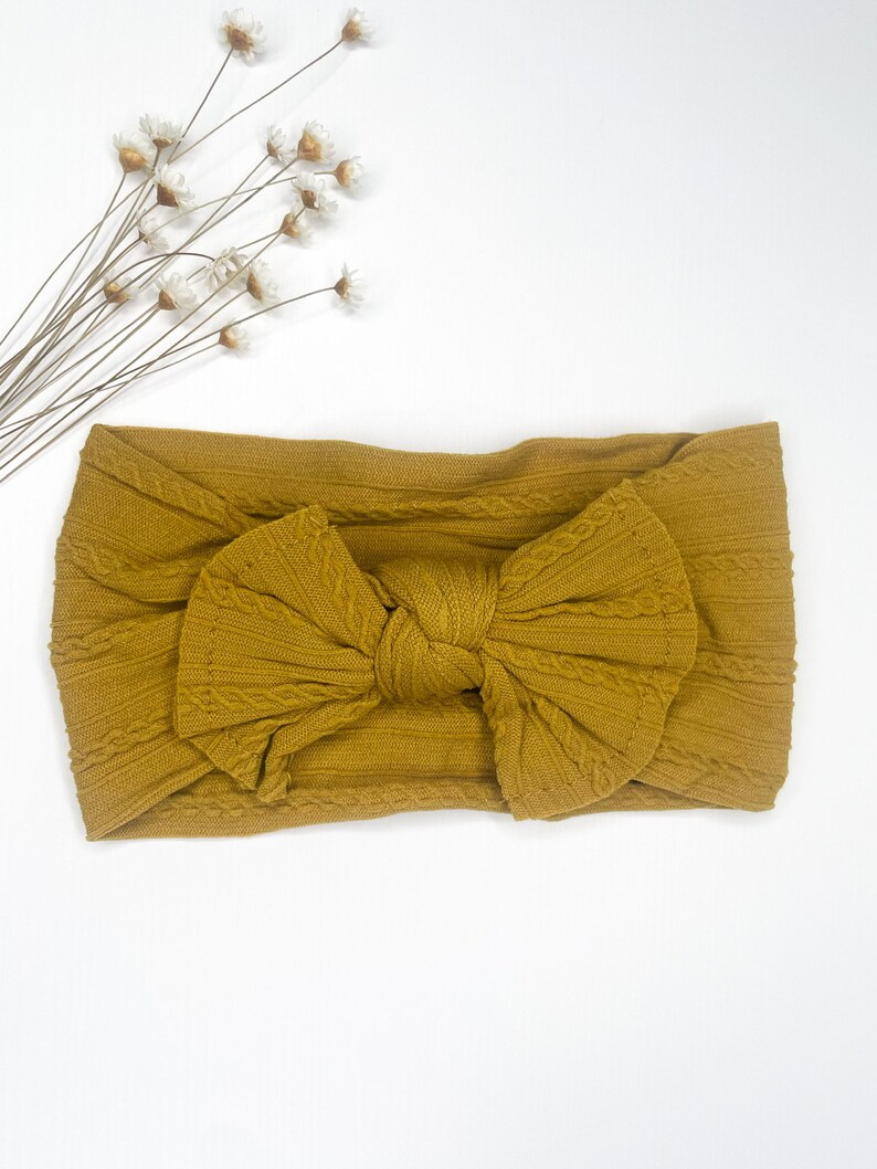 May include: A mustard yellow knotted headband with a cable knit texture. The headband is made of soft, stretchy fabric and is perfect for adding a touch of style to any outfit.