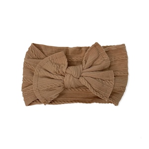 May include: A brown cable knit headband with a large bow. The headband is made of a soft, stretchy fabric.