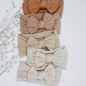 May include: Five different hair bows in various shades of brown and beige. The bows are made of a soft, textured fabric and have a knotted center. The bows are arranged in a vertical stack, with the darkest bow at the top and the lightest bow at the bottom.