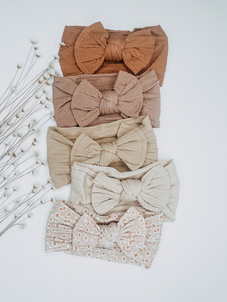 May include: Five different hair bows in various shades of brown and beige. The bows are made of a soft, textured fabric and have a knotted center. The bows are arranged in a vertical stack, with the darkest bow at the top and the lightest bow at the bottom.