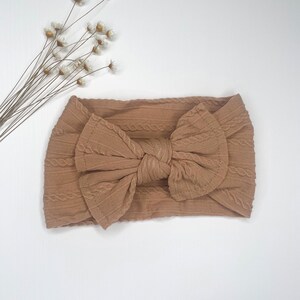 May include: A brown cable knit headband with a large bow. The headband is made of a soft, stretchy fabric.