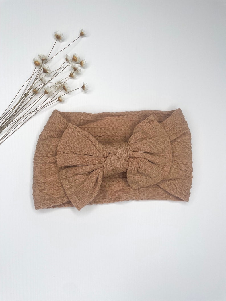 May include: A brown cable knit headband with a large bow. The headband is made of a soft, stretchy fabric.