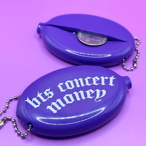 May include: Two purple coin purses with a silver chain. One purse has the text "bts concert money" printed on it in white. The other purse is open and has a coin inside.