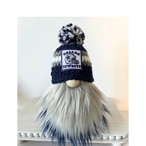 May include: A Dallas Cowboys gnome with a blue, white, and gray striped hat with a pom-pom on top. The hat has a Dallas Cowboys logo patch on the front. The gnome has a long, white and gray beard.