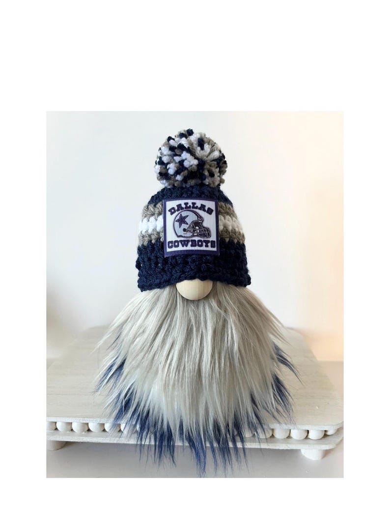 May include: A Dallas Cowboys gnome with a blue, white, and gray striped hat with a pom-pom on top. The hat has a Dallas Cowboys logo patch on the front. The gnome has a long, white and gray beard.