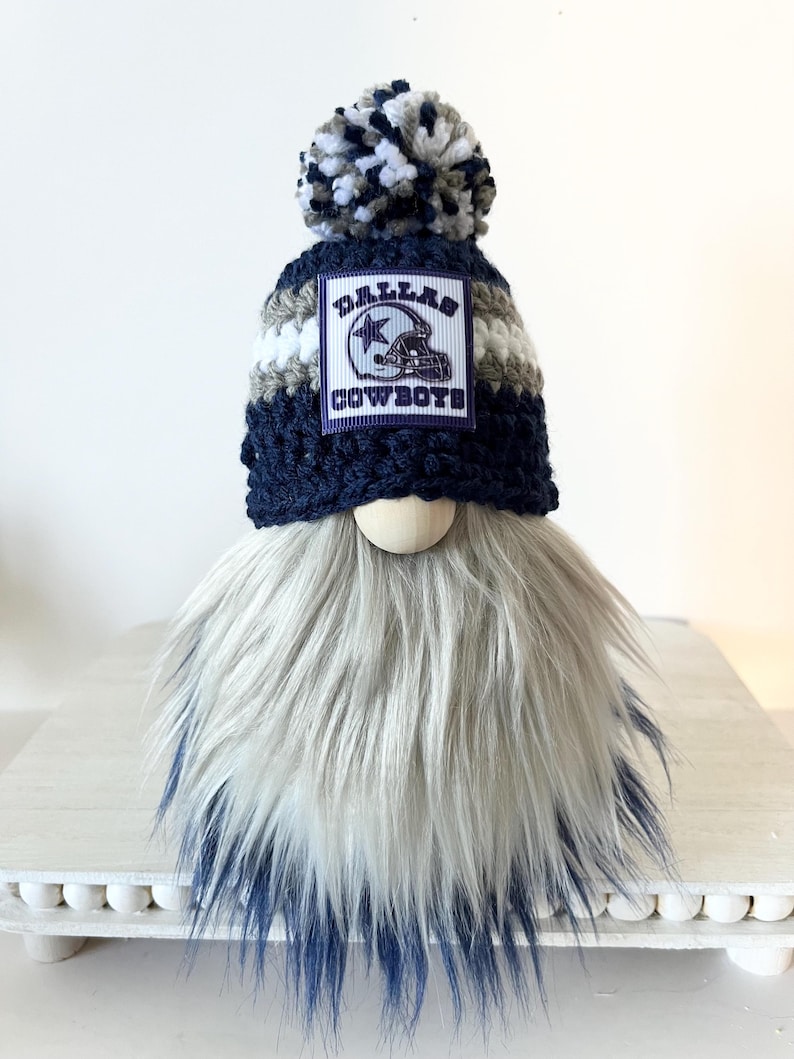 May include: A crocheted gnome with a blue and white pom-pom hat. The hat has a white and gray striped band and a patch that says "Dallas Cowboys". The gnome has a long, white and blue beard.
