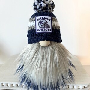 May include: A gnome with a white and blue striped hat with a Dallas Cowboys logo patch and a white, blue, and gray pom pom. The gnome has a long, white and gray beard with a blue ombre effect.