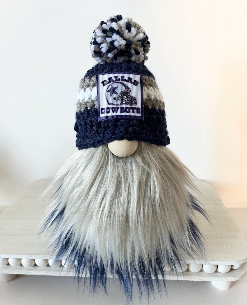 May include: A gnome with a white and blue striped hat with a Dallas Cowboys logo patch and a white, blue, and gray pom pom. The gnome has a long, white and gray beard with a blue ombre effect.