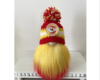 May include: A decorative gnome featuring a long yellow beard with red tips, wearing a red, white, and yellow crocheted hat topped with a matching pom-pom. The hat displays a circular Kansas City Chiefs logo patch. The text Little Sky Creation SF is visible.