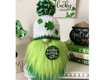 May include: A green and white gnome wearing a white crocheted hat with green shamrocks and a green pom-pom. The gnome is holding a round sign that says "Happy St. Patrick's Day".