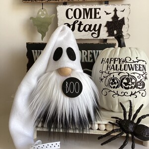 May include: A white ghost gnome with a black fur beard and a black circle with the word "BOO" on it. The gnome is sitting on a white shelf with a white pumpkin with black lettering that says "Happy Halloween" and a black spider.