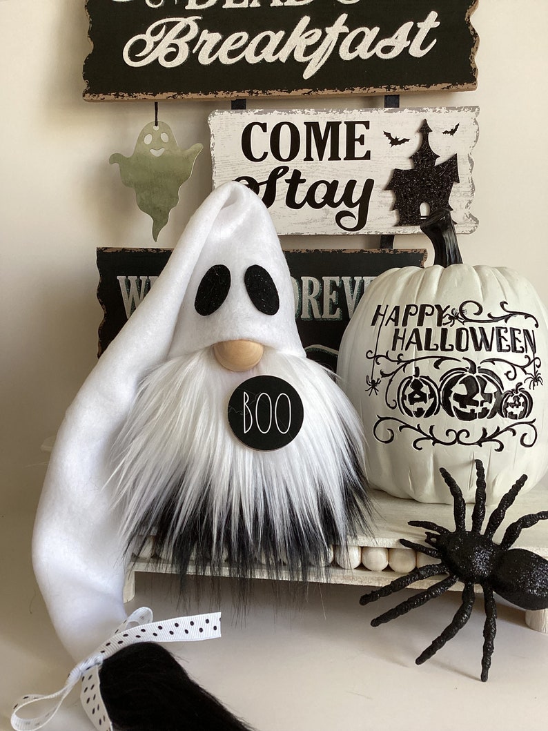 May include: A white ghost gnome with a black fur beard and a black circle with the word "BOO" on it. The gnome is sitting on a white shelf with a white pumpkin with black lettering that says "Happy Halloween" and a black spider.