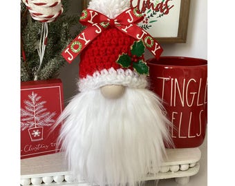 May include: A Christmas gnome with a red and white knitted hat, a long white beard, and a wooden nose. The hat has a ribbon that says "JOY" and holly berries. A red mug with "Tingle Bells" is next to the gnome.