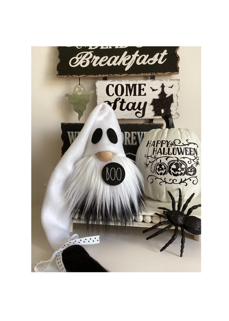 May include: A white ghost gnome with a black and white fur beard and a black circle with the word "BOO" on it. The gnome is sitting on a white shelf with a white pumpkin with black lettering that says "Happy Halloween" and a black spider.