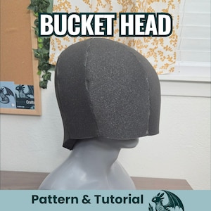 May include: A dark grey foam head covering with a rounded top and straight sides, displayed on a grey mannequin head. The text BUCKET HEAD is printed in white letters above the headpiece, and Pattern & Tutorial is printed at the bottom.