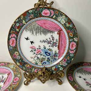May include: Three porcelain plates with a floral design. The plates feature a pink and white floral pattern with birds and a pink curtain. The plates are on a gold metal stand.