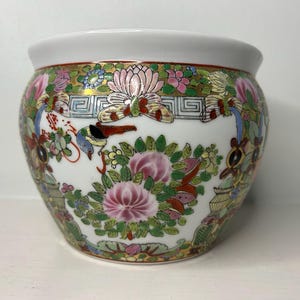 May include: A white ceramic bowl featuring a colorful hand-painted design of pink lotus flowers, green foliage, and butterflies. The rim is accented with a gold-toned border and a geometric pattern, while the body displays intricate floral motifs.