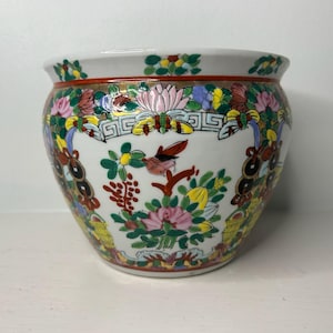 May include: A white ceramic bowl featuring a colorful hand-painted floral and butterfly pattern. The exterior displays intricate designs in pink, green, yellow, and red, with a decorative border around the rim and a wide, rounded body shape.