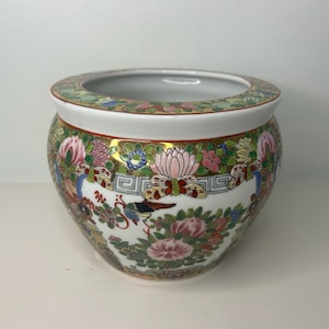 May include: A round porcelain planter featuring a white base decorated with colorful floral patterns, butterflies, and gold-toned accents. The bowl has a wide, open top with a patterned rim and a curved body that tapers toward the base.