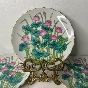 May include: A set of three white porcelain plates with scalloped edges and gold trim, each featuring a vibrant illustration of pink lotus flowers, green lily pads, and yellow reeds. One plate is displayed on an ornate brass metal stand.