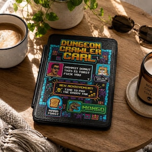 May include: A black e-reader cover featuring pixel art graphics and the text Dungeon Crawler Carl, Dammit Donut This Is Toast Fuck You, This Is An Outrage, New Achievement! Time To Pay The Daddy Tax, Mama Toast, and Mongo with a dinosaur icon.