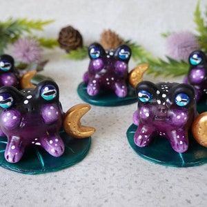 May include: Five purple and black frog figurines with white spots and gold crescent moons. Each frog is sitting on a green lily pad.