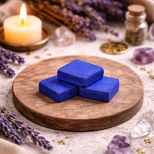 Può includere: Tre oggetti quadrati blu cobalto sono impilati su un vassoio rotondo di legno. Una candela accesa in un portacandele di ottone, lavanda secca, cristalli e piccole stelle dorate circondano il vassoio, creando un'estetica mistica.