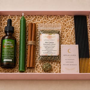 Puede incluir: Una caja rosa llena de artículos rituales. Incluye una botella de "Money Drawing Ritual Oil", una vela verde, palitos de canela, una bolsa de hierbas, varillas de incienso y una guía ritual. También hay una vela encendida y un cristal.