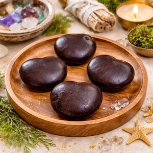 May include: A wooden plate holds four dark brown, heart-shaped objects. The plate is surrounded by crystals, a lit candle, and other spiritual items. The objects appear to be smooth and have a slightly glossy finish.