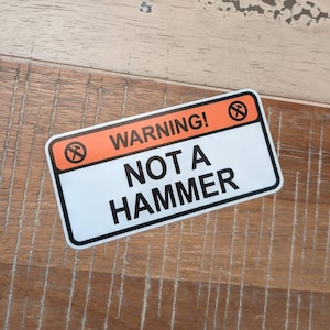May include: A white warning sign with a red and orange border. The sign has a black hammer with a red circle and diagonal line through it. The text on the sign reads "WARNING! NOT A HAMMER".