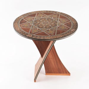 May include: A round wooden table with a geometric inlay design in shades of brown, gold, and black. The table has a unique X-shaped base.